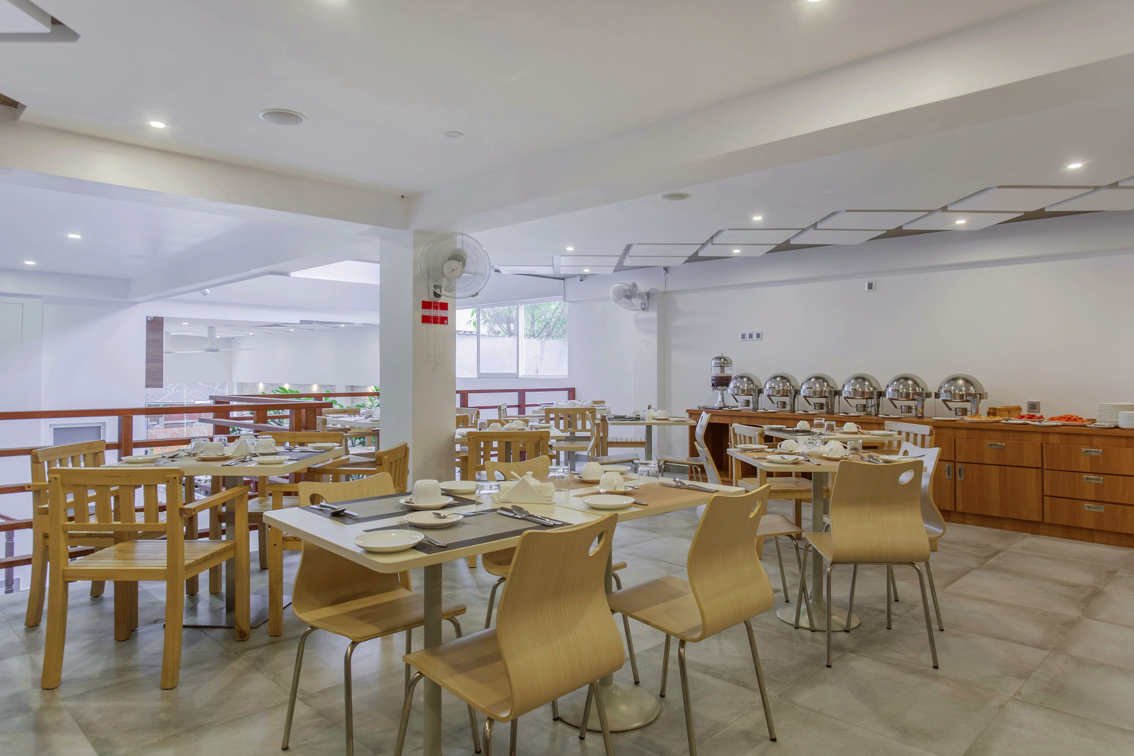 A spacious dining area with multiple tables and chairs, a kitchen area with buffet setup, and a view of the outdoors.