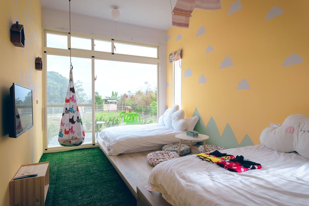 A room with two beds, a green chair, a hanging bag, and a small table.
