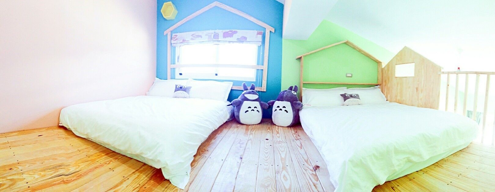 A room with two beds, wooden flooring, a window with a blue wall, and a small wooden house structure.