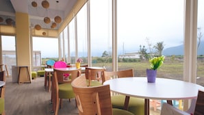 A dining area with wooden chairs and tables, large windows with a view of the outdoors, and a ceiling with hanging lights.