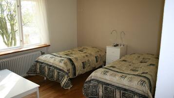 Une chambre avec deux lits, une fenêtre avec des rideaux, un radiateur, une table blanche et une table de chevet avec une lampe.