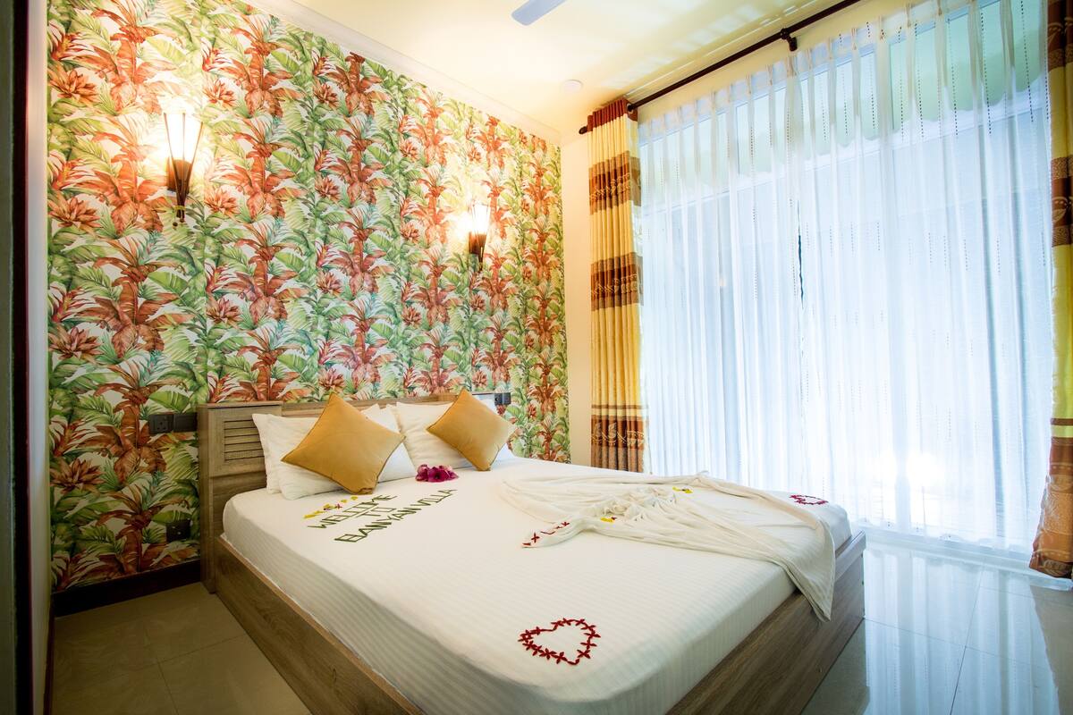 Una habitación de hotel con papel tapiz floral, una cama bien hecha con un adorno con forma de corazón y una ventana con cortinas de gasa.