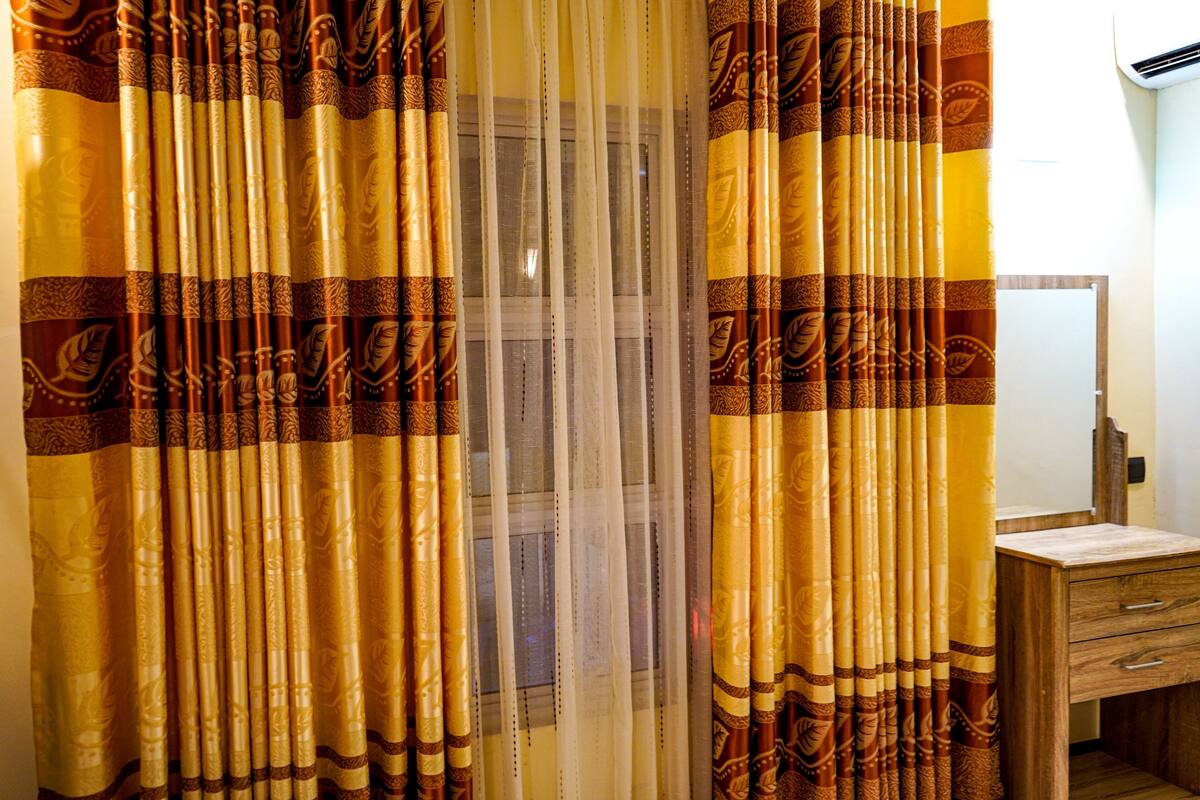 Una habitación con ventana cubierta por cortinas estampadas, una mesita de noche de madera y un espejo.
