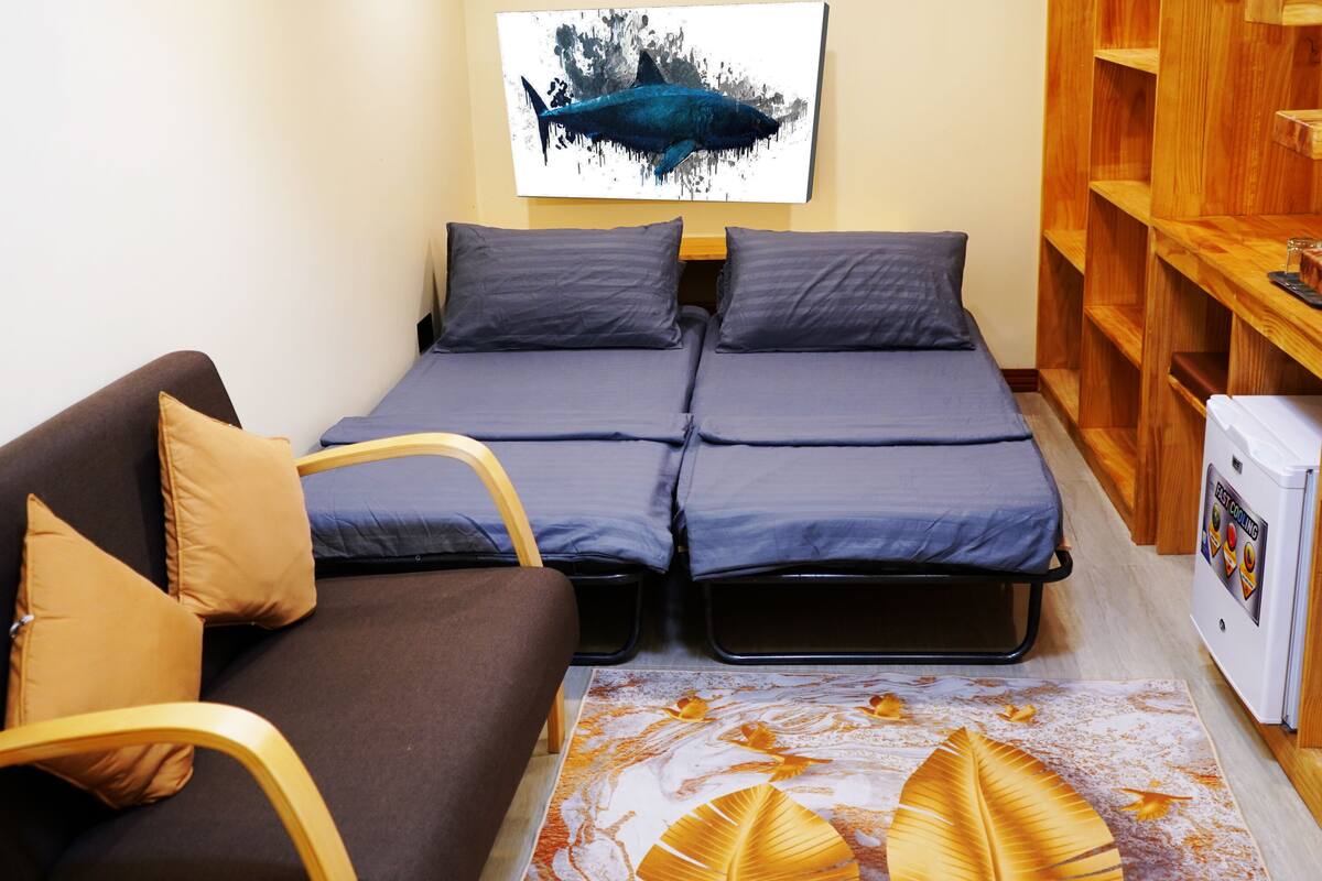 Una habitación pequeña con sofá, cama con colchón en el suelo, estantería de madera y refrigerador.