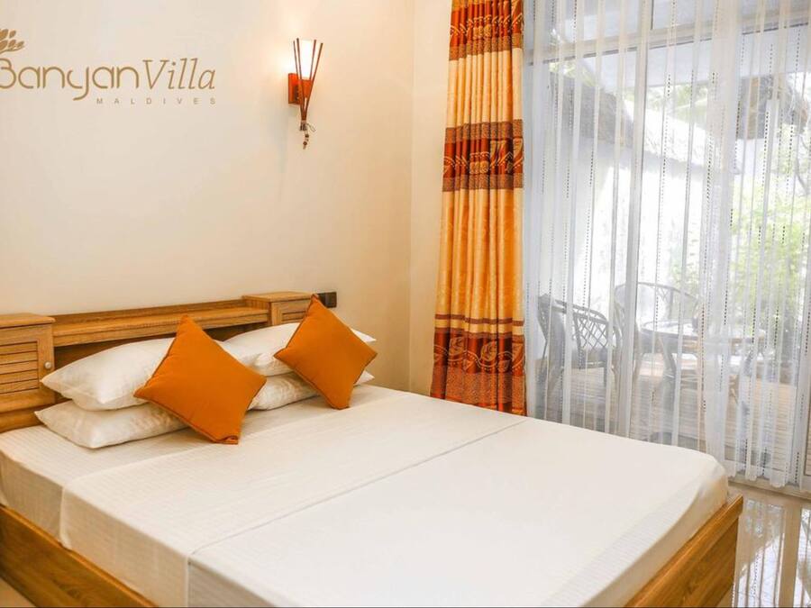 A bedroom with a wooden bed, white bedding, and orange pillows. There is a wall-mounted light fixture and a window with curtains.