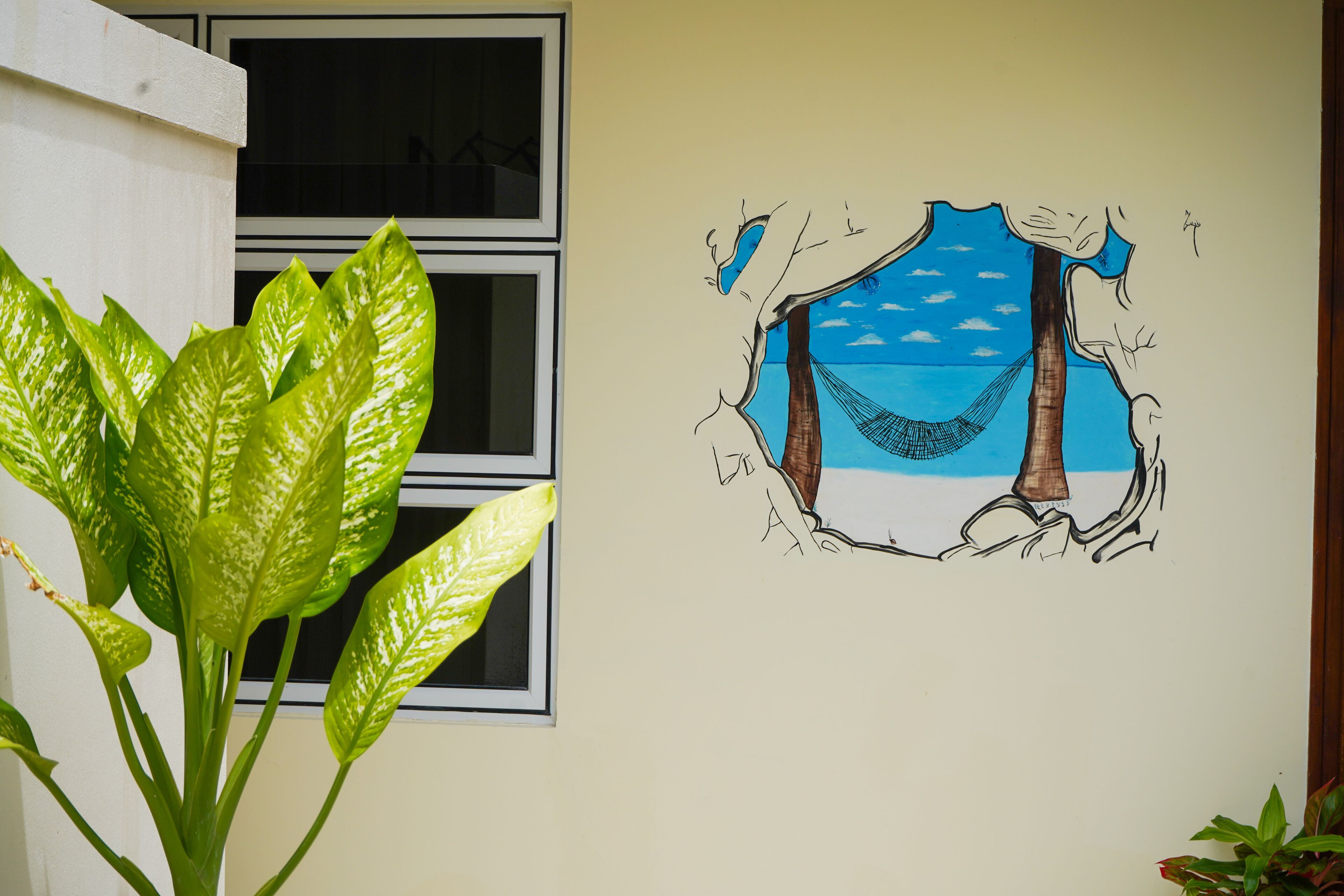 A wall with a large green plant and a mural of a hammock between two trees overlooking a beach.