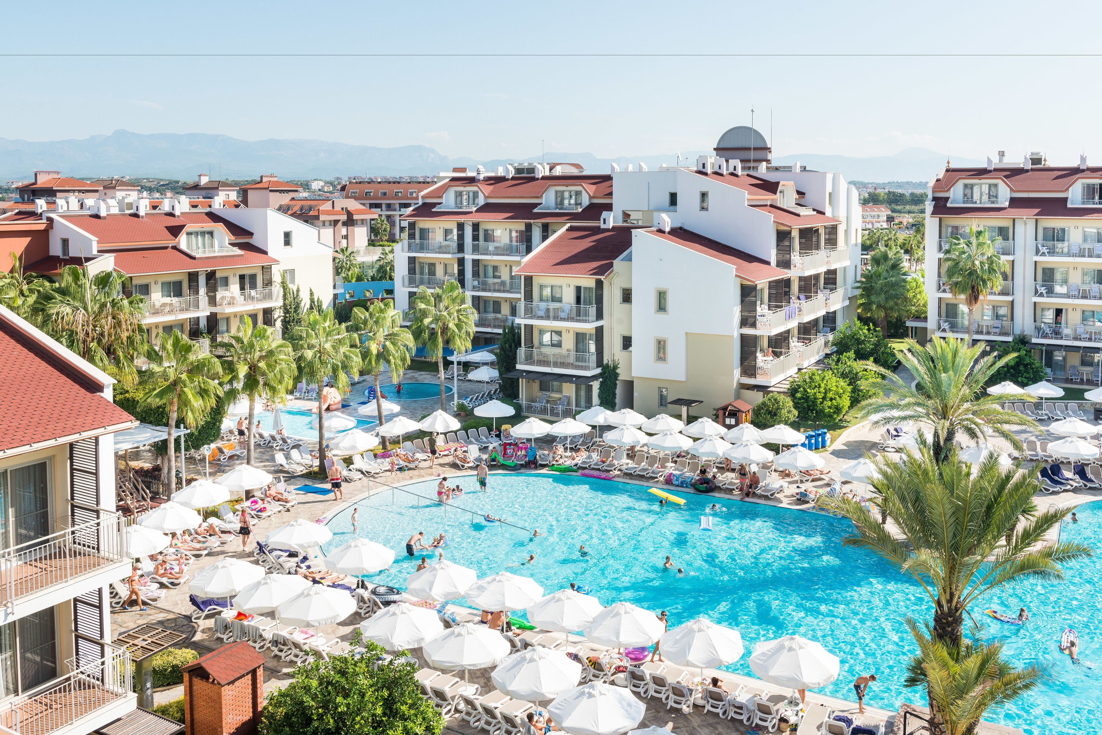 Un resort con una amplia piscina rodeada de tumbonas y sombrillas, varios balcones y vistas a montañas distantes.