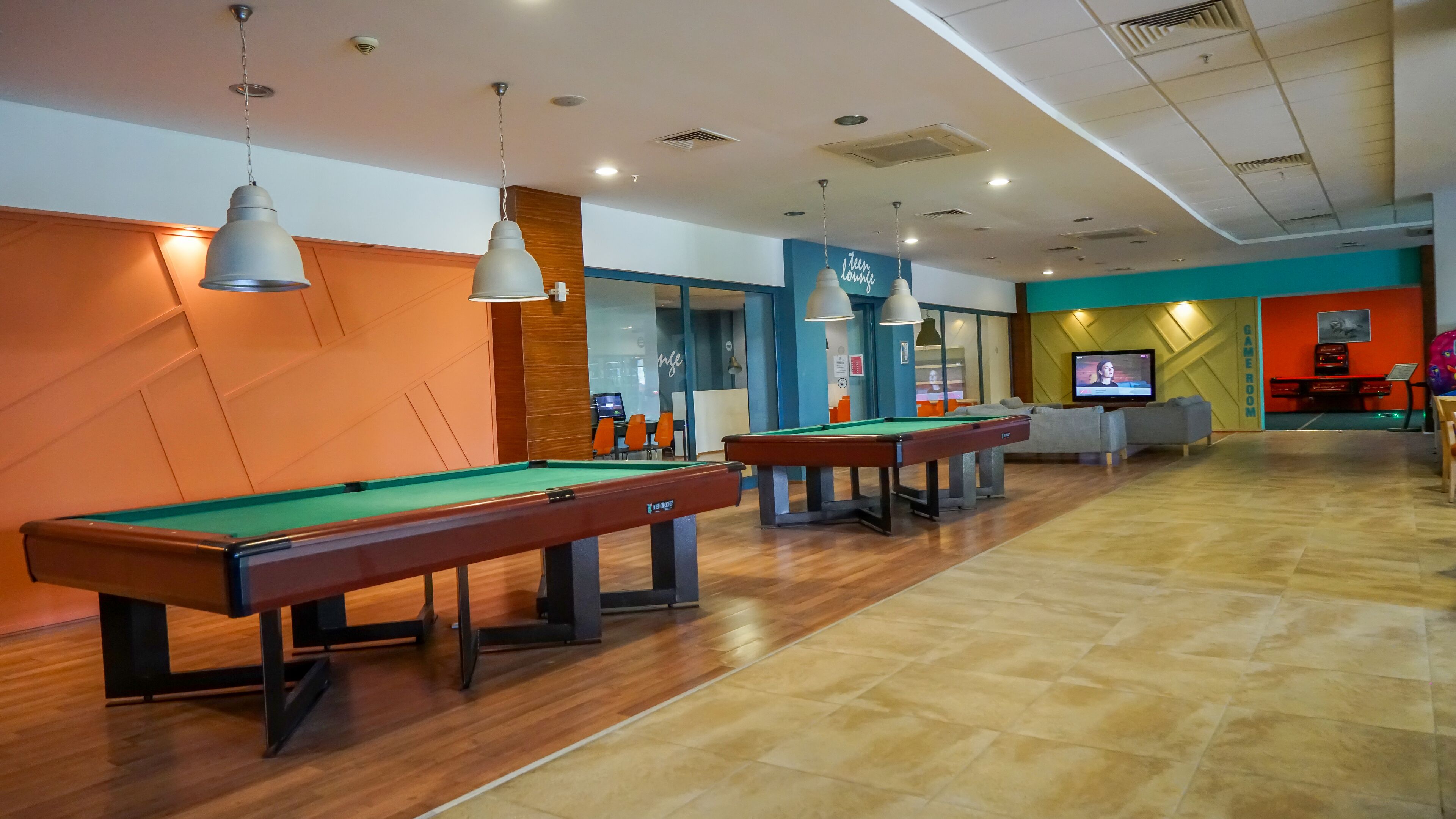 A pool hall with multiple pool tables, overhead lighting, and a television screen in the background.
