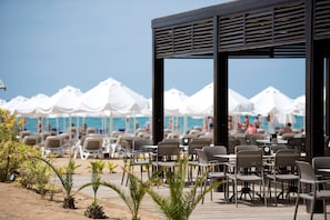 Un restaurante frente a la playa con mesas al aire libre, sombrillas blancas y vistas a la playa y al océano.