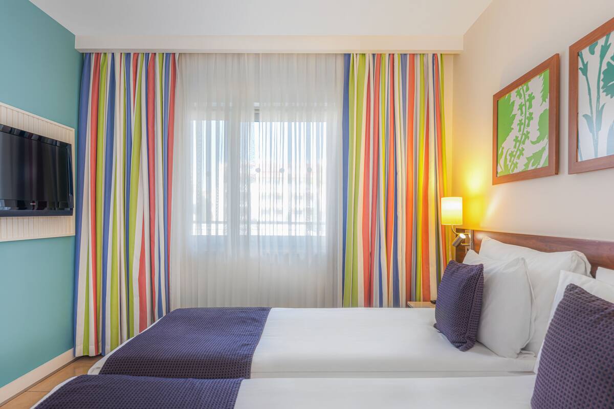A hotel room with two beds, a TV, a window with colorful curtains, and a lamp.
