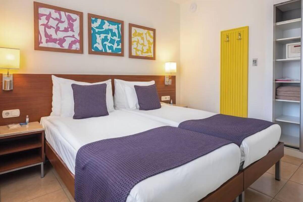 A double bed with white and purple bedding, a nightstand with a lamp, and abstract art on the wall.