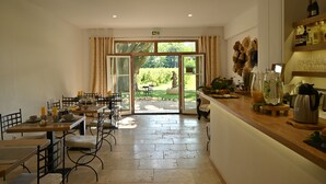 Comedor con mesas y sillas, mostrador de cocina con distintos objetos, y vista al jardín a través de puertas de vidrio.