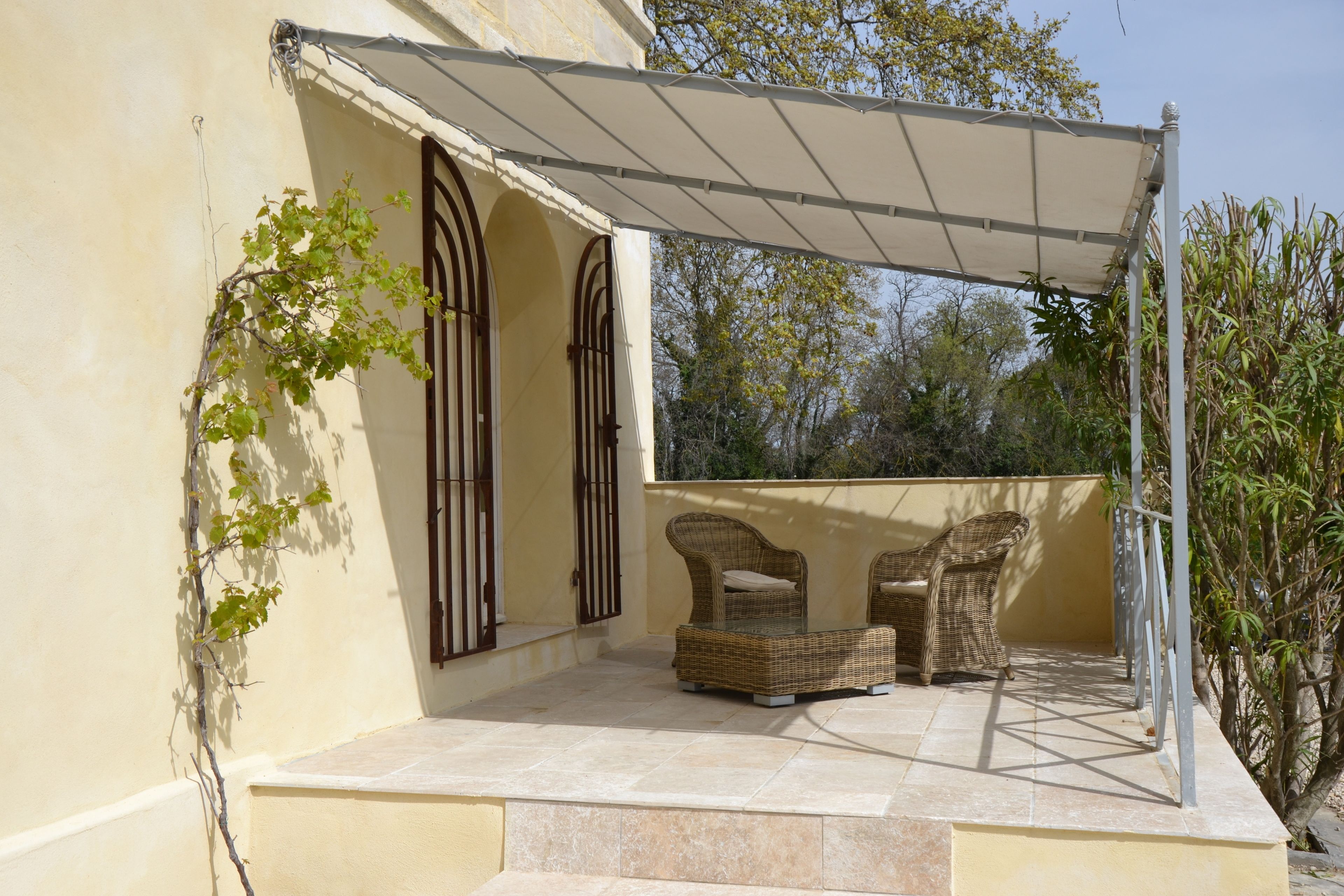 Un espacio de estar al aire libre techado con mobiliario de mimbre, una pérgola y vistas a árboles y cielo.