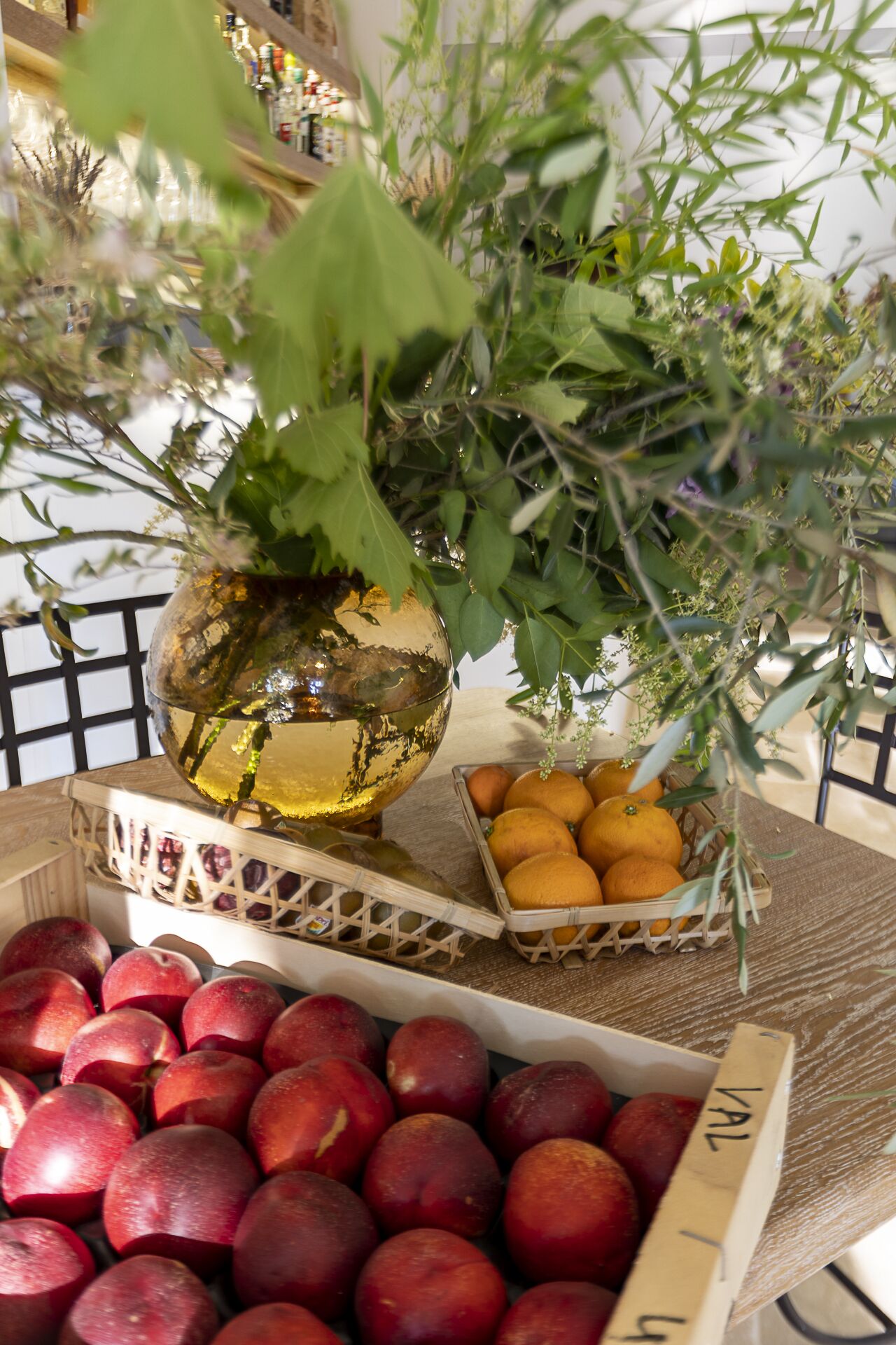 A table with a decorative glass vase, a basket of oranges, and a crate of red apples.