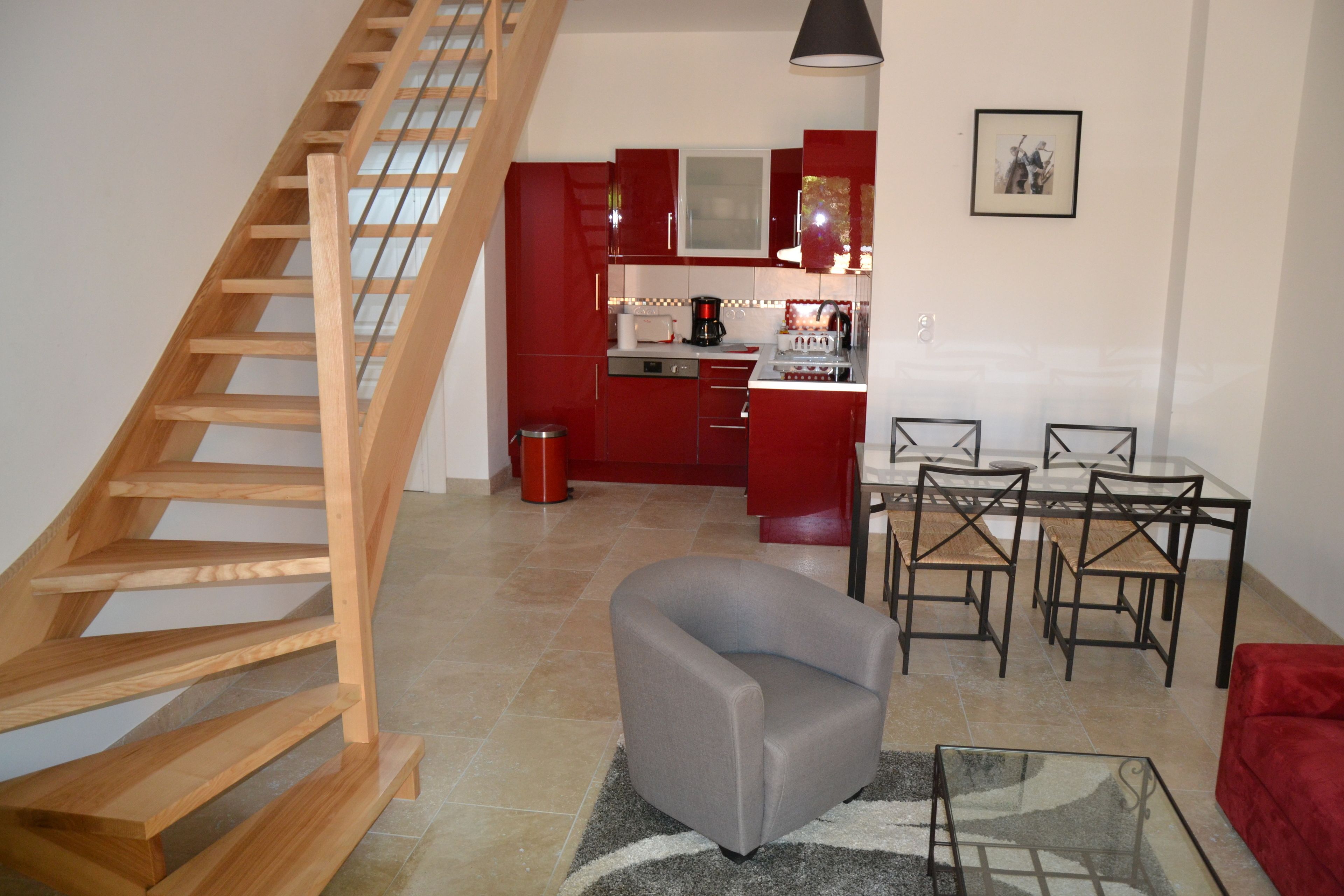 A modern apartment with a wooden staircase, a grey armchair, a dining area with chairs, and a kitchen with red cabinets.