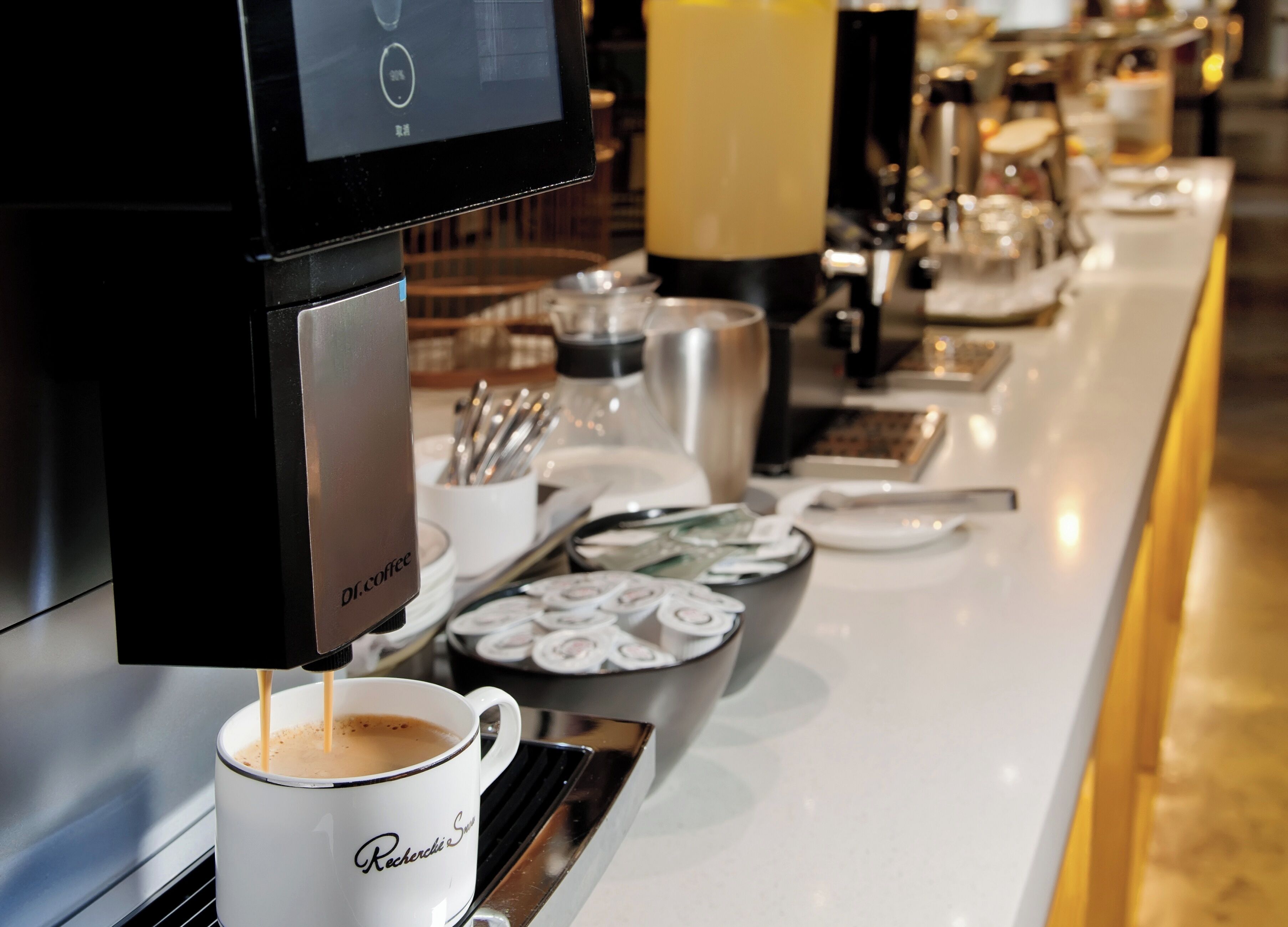 A coffee machine dispensing coffee into a cup, with a tray of pastries and a coffee station in the background.