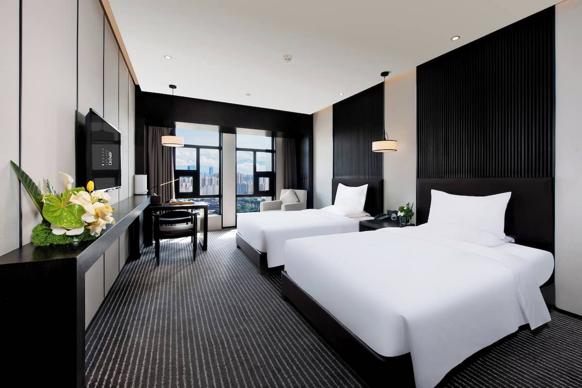 A modern hotel room with two beds, a desk, and a large window offering a city view.