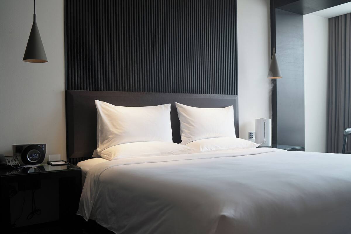 A modern hotel room with a large bed, a bedside table with a lamp, and a wall-mounted panel.