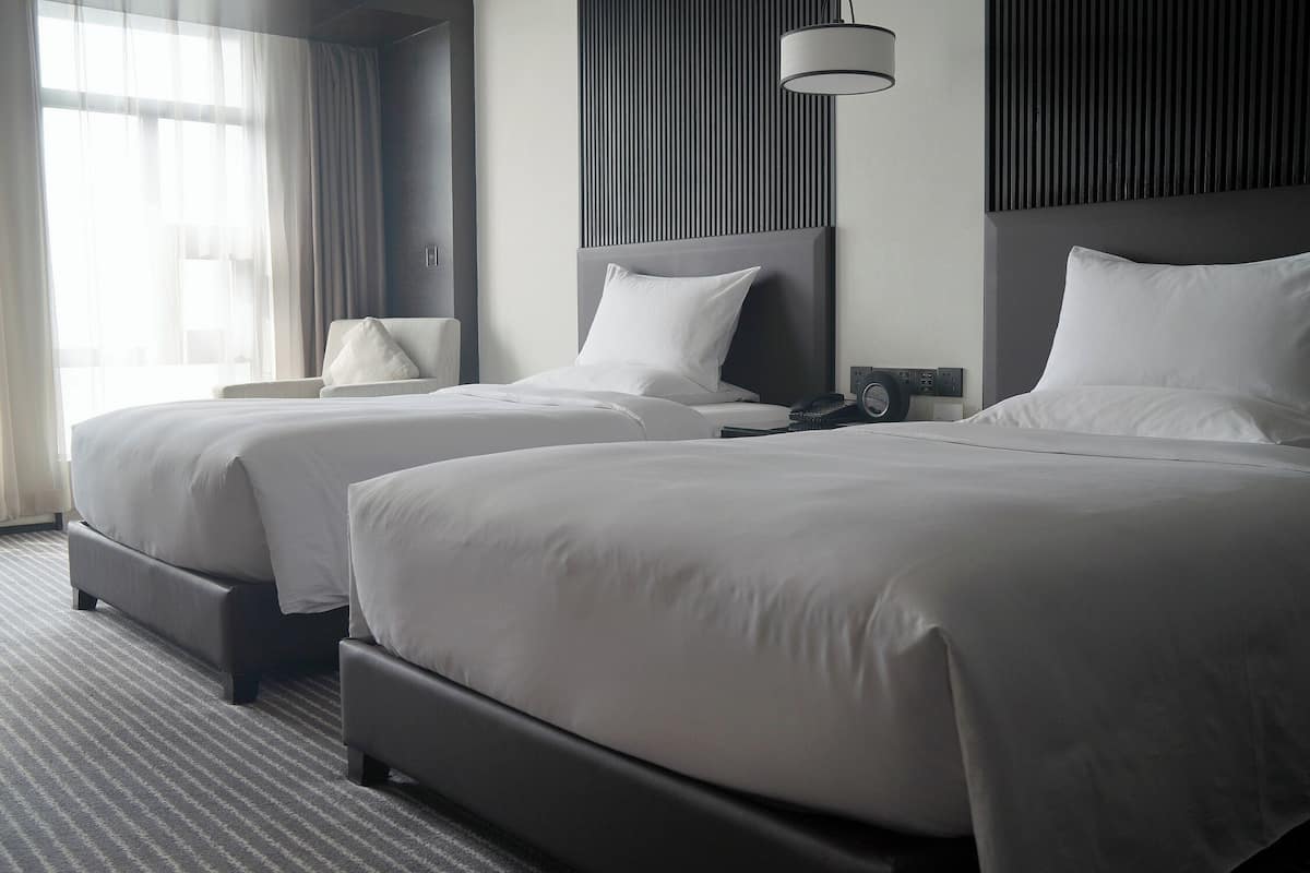 A hotel room with two beds, a large window with sheer curtains, a bedside table with a lamp, and a modern wall panel.