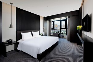 Ein modernes Hotelzimmer mit einem großen Bett, einem Schreibtisch, einem Sessel, einem Fernseher und einem Fenster mit Blick auf die Stadt.