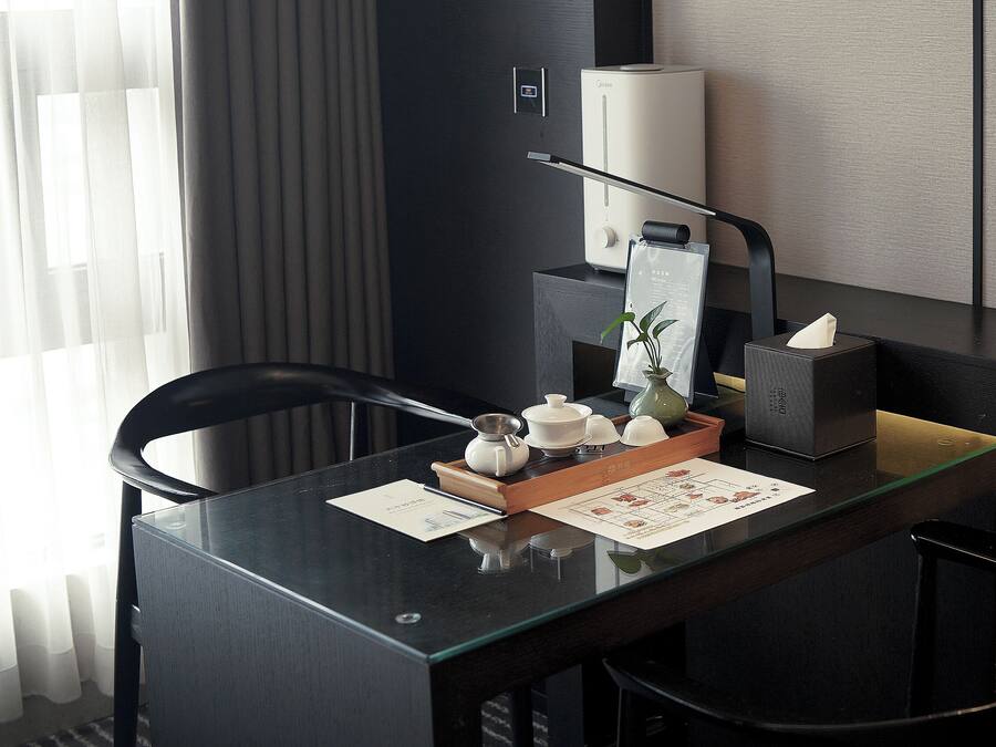 Habitación de hotel moderna con un escritorio de madera oscura, un juego de té, una planta y una lámpara.