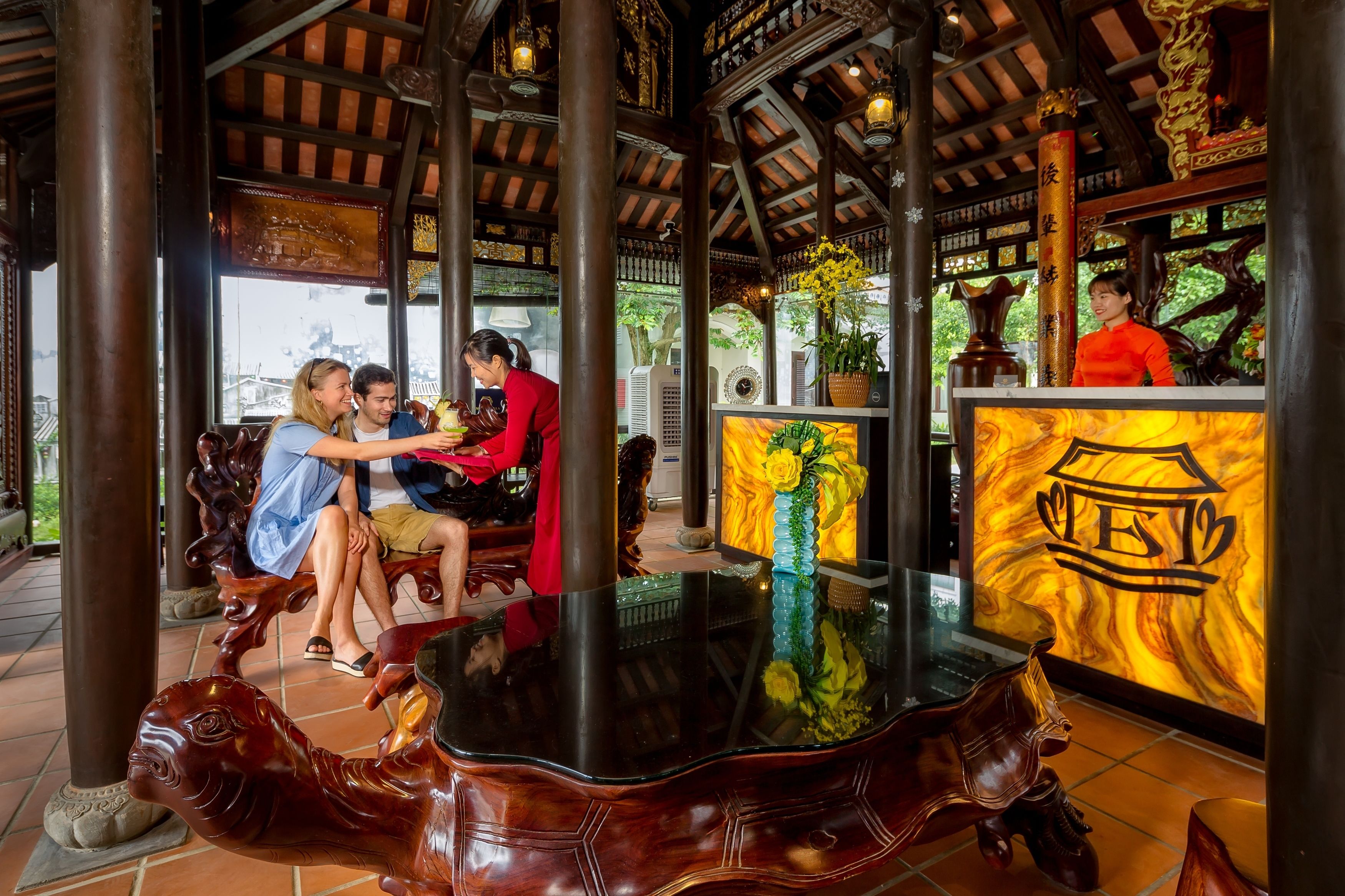 Een klassiek houten paviljoen met een glazen tafel in het midden, omringd door sierlijke houtsnijwerken en zuilen.