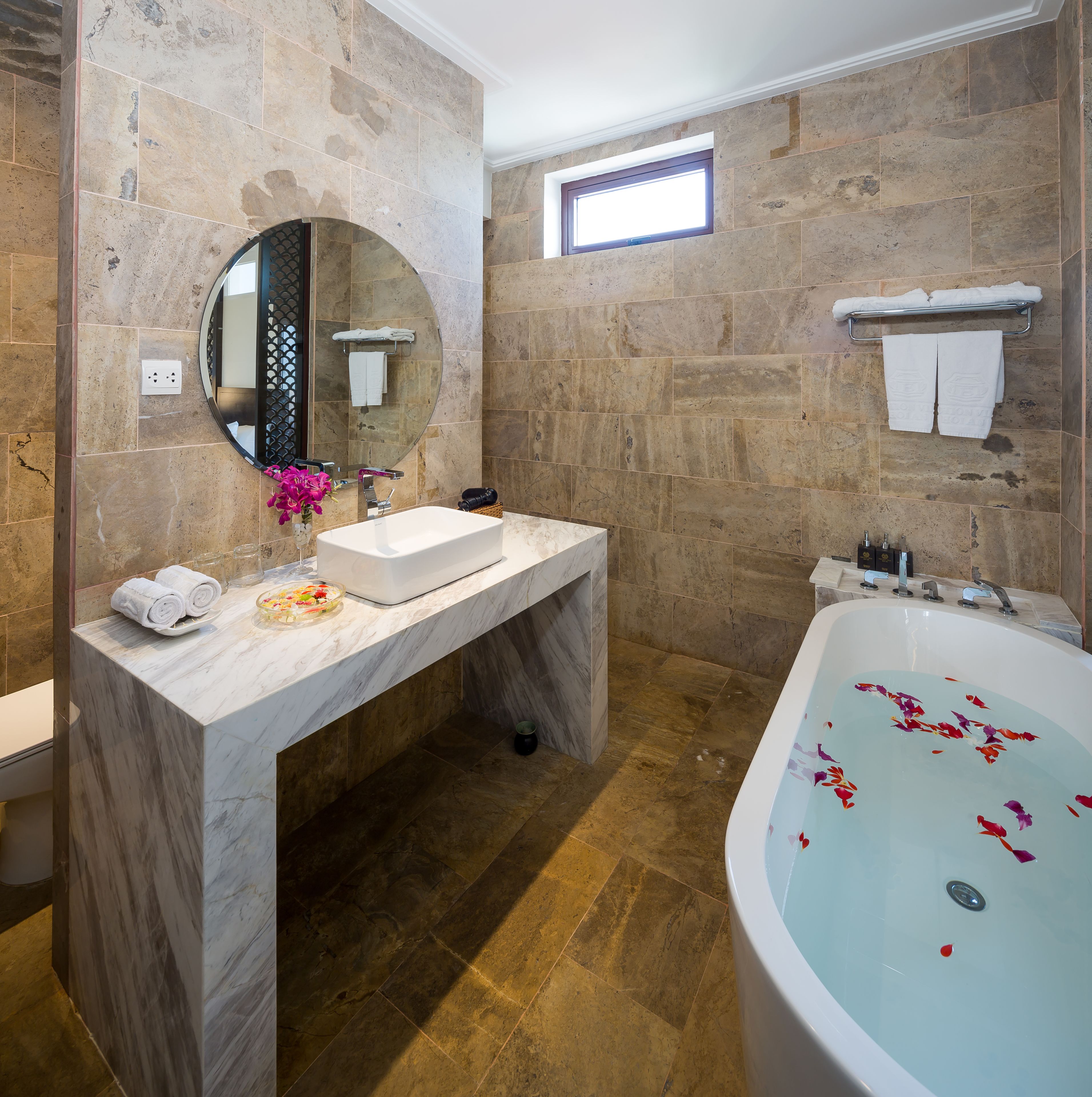 Un baño con tocador de mármol, una bañera independiente con pétalos de rosa, una ventana y un perchero para toallas.