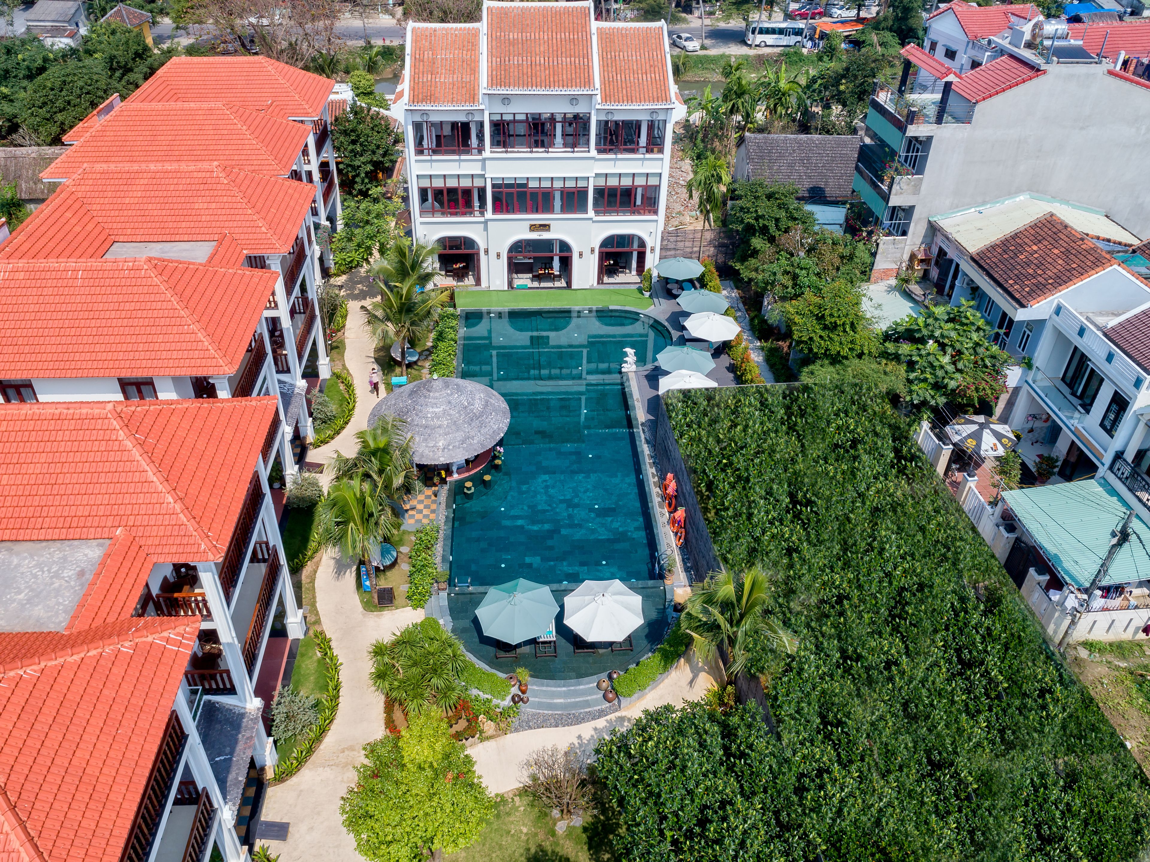 An aerial view of a resort with a swimming pool, multiple buildings with red roofs, and lush greenery.