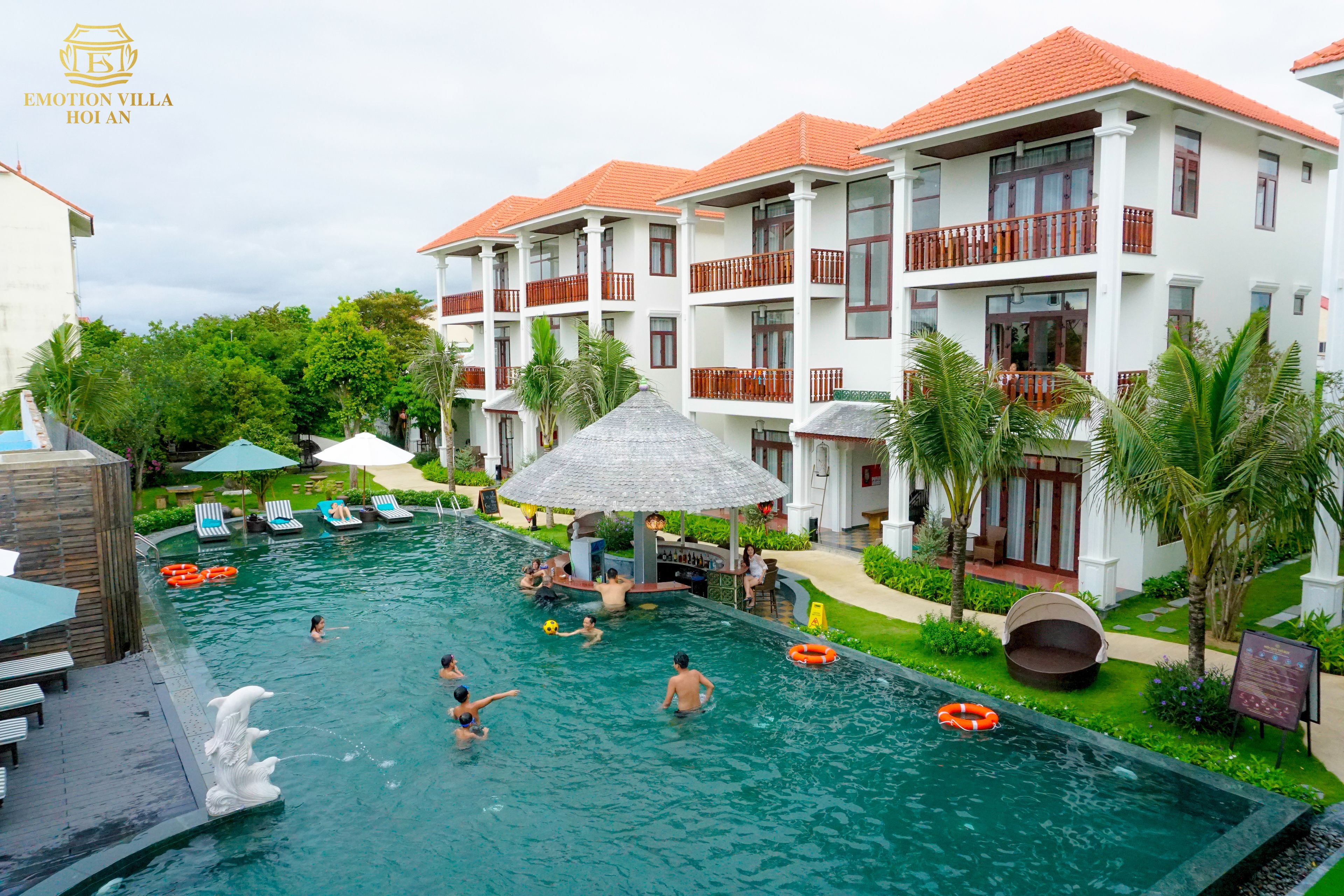 A swimming pool area with multiple levels of buildings, lounge chairs, and a thatched-roof structure.
