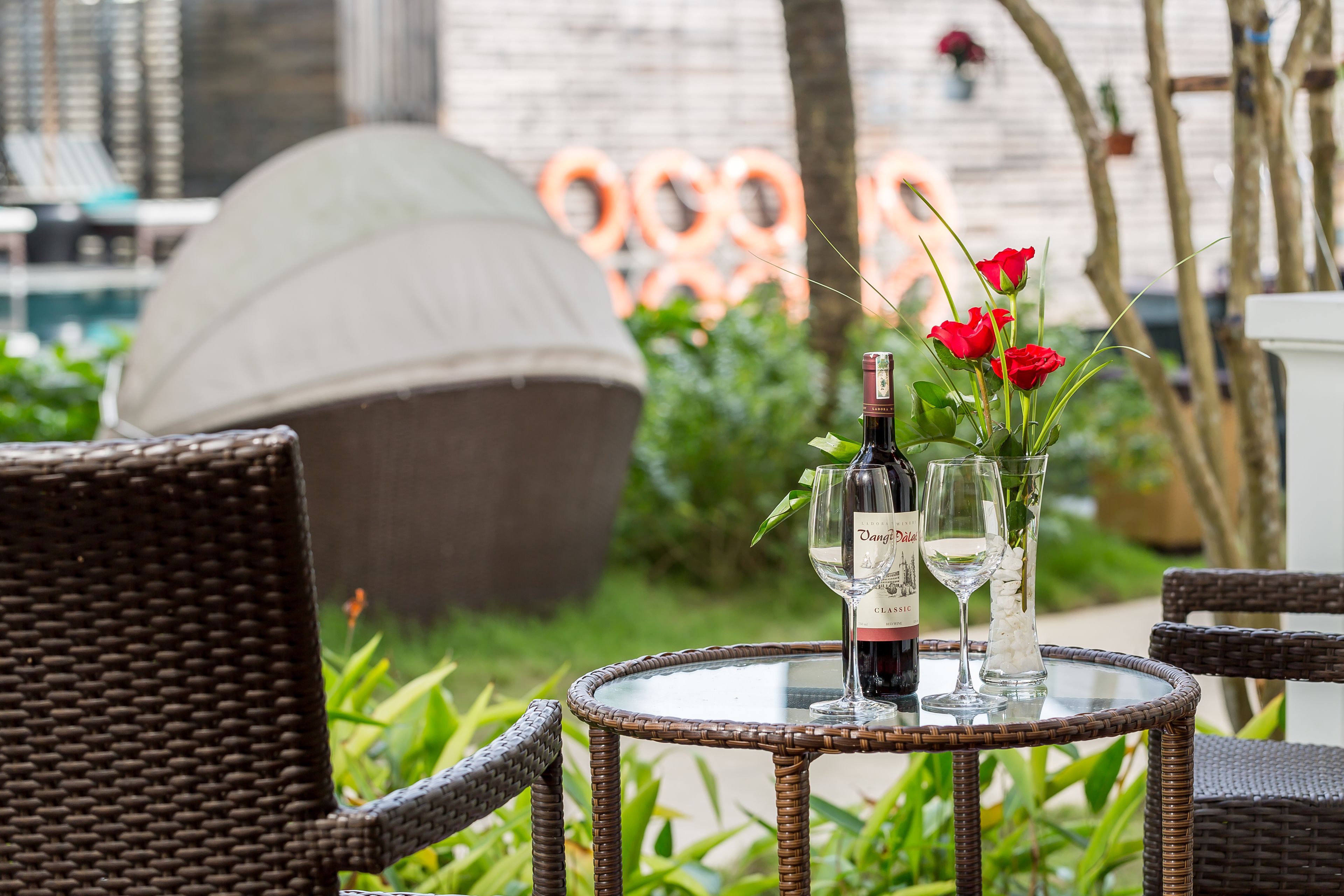 A wicker table with a bottle of wine and two wine glasses, surrounded by greenery and red roses.