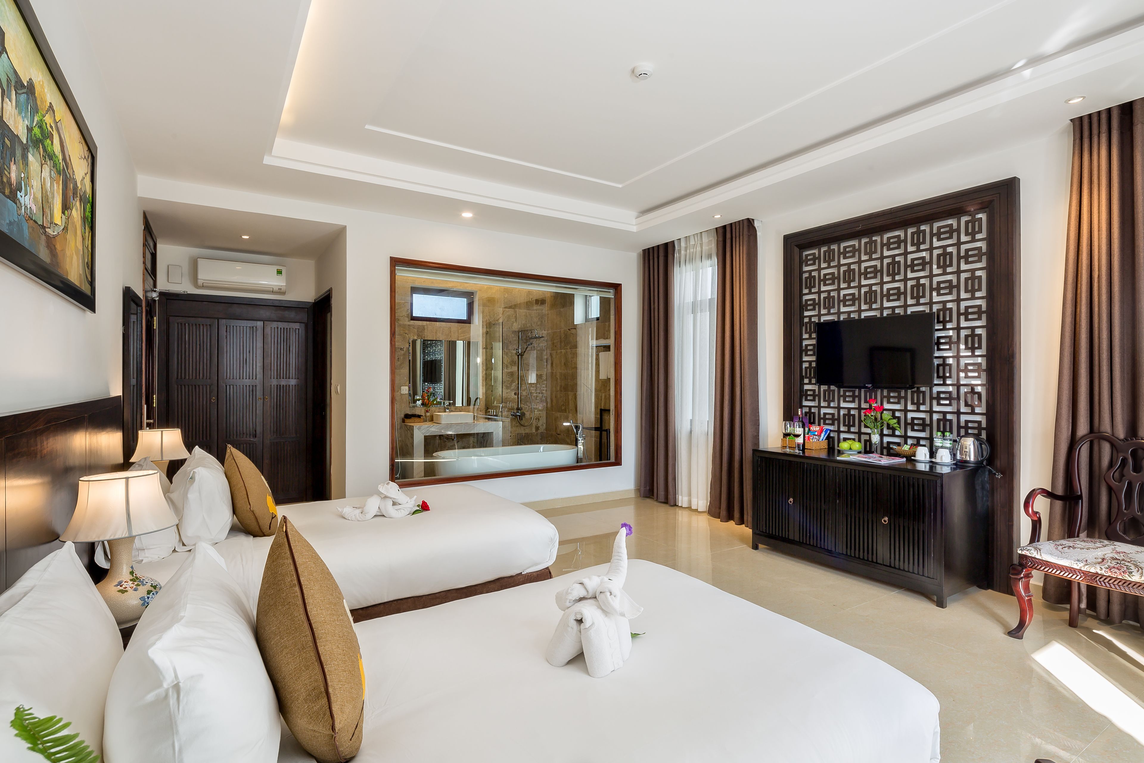 A hotel room with a large bed, a sitting area, a TV, a minibar, and a bathroom visible through a glass door.