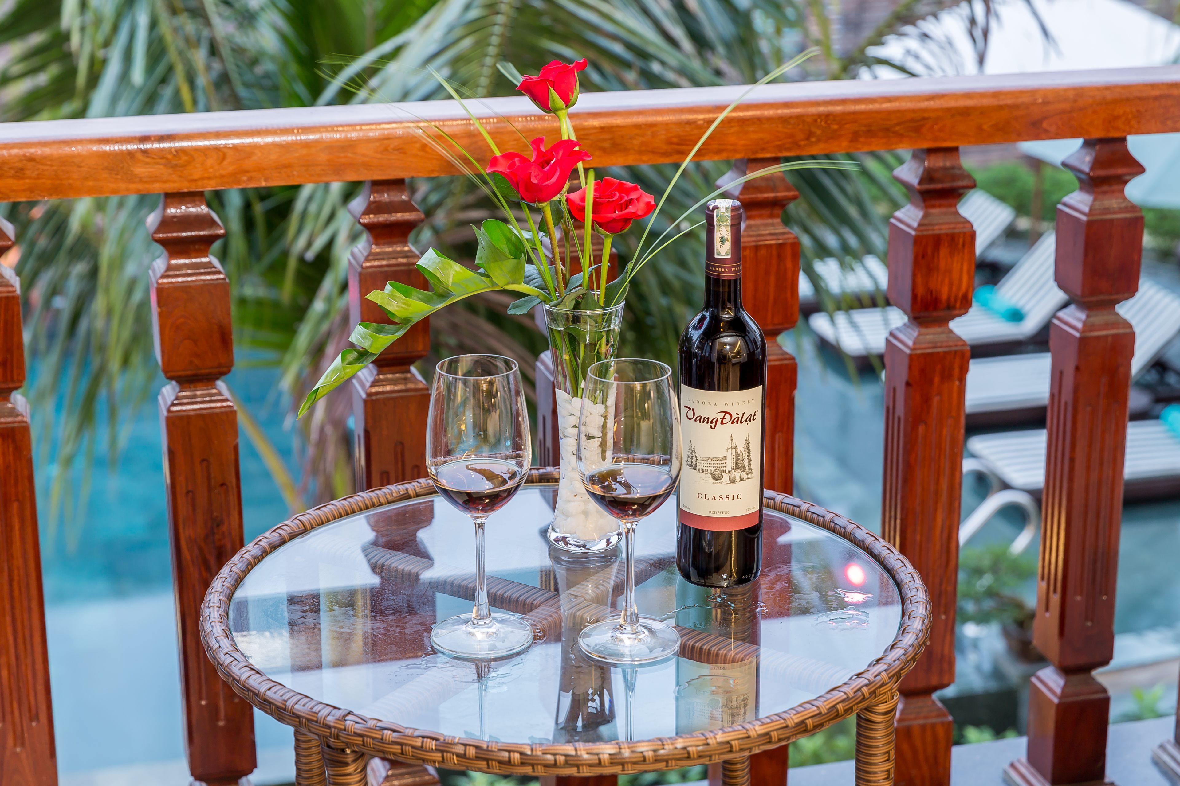 Una mesa de cristal con una botella de vino, dos copas de vino y un florero con rosas rojas en un balcón.