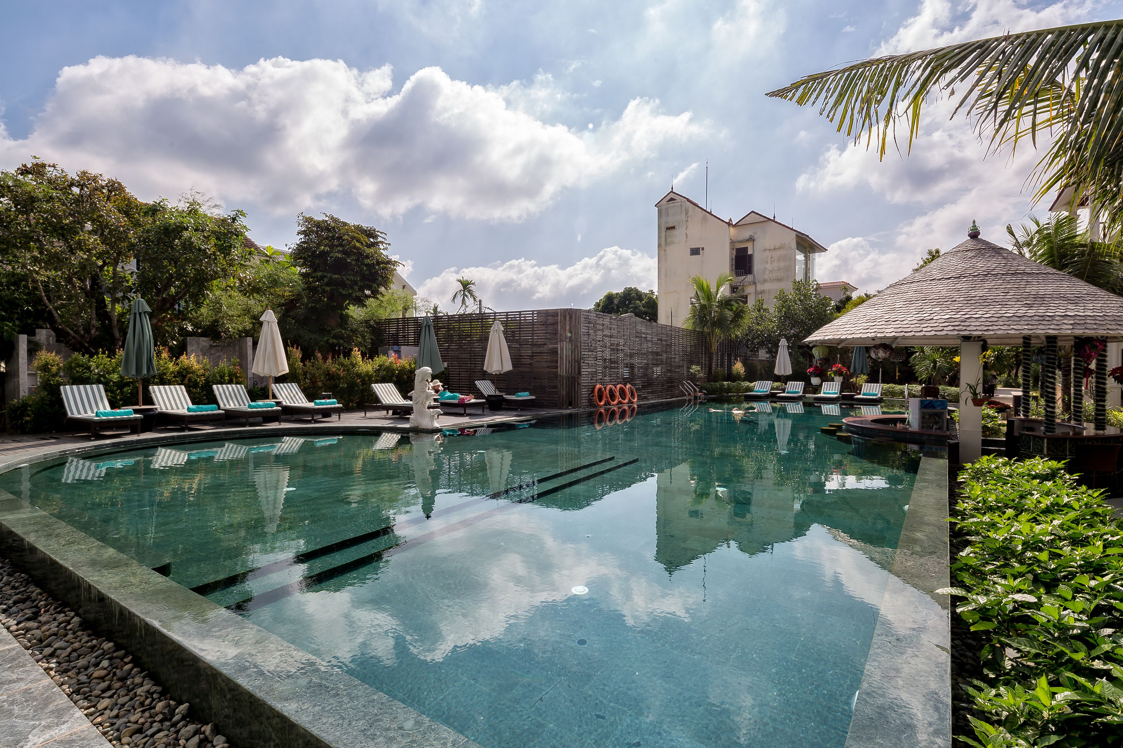 A swimming pool with lounge chairs and umbrellas, surrounded by greenery and a building in the background.