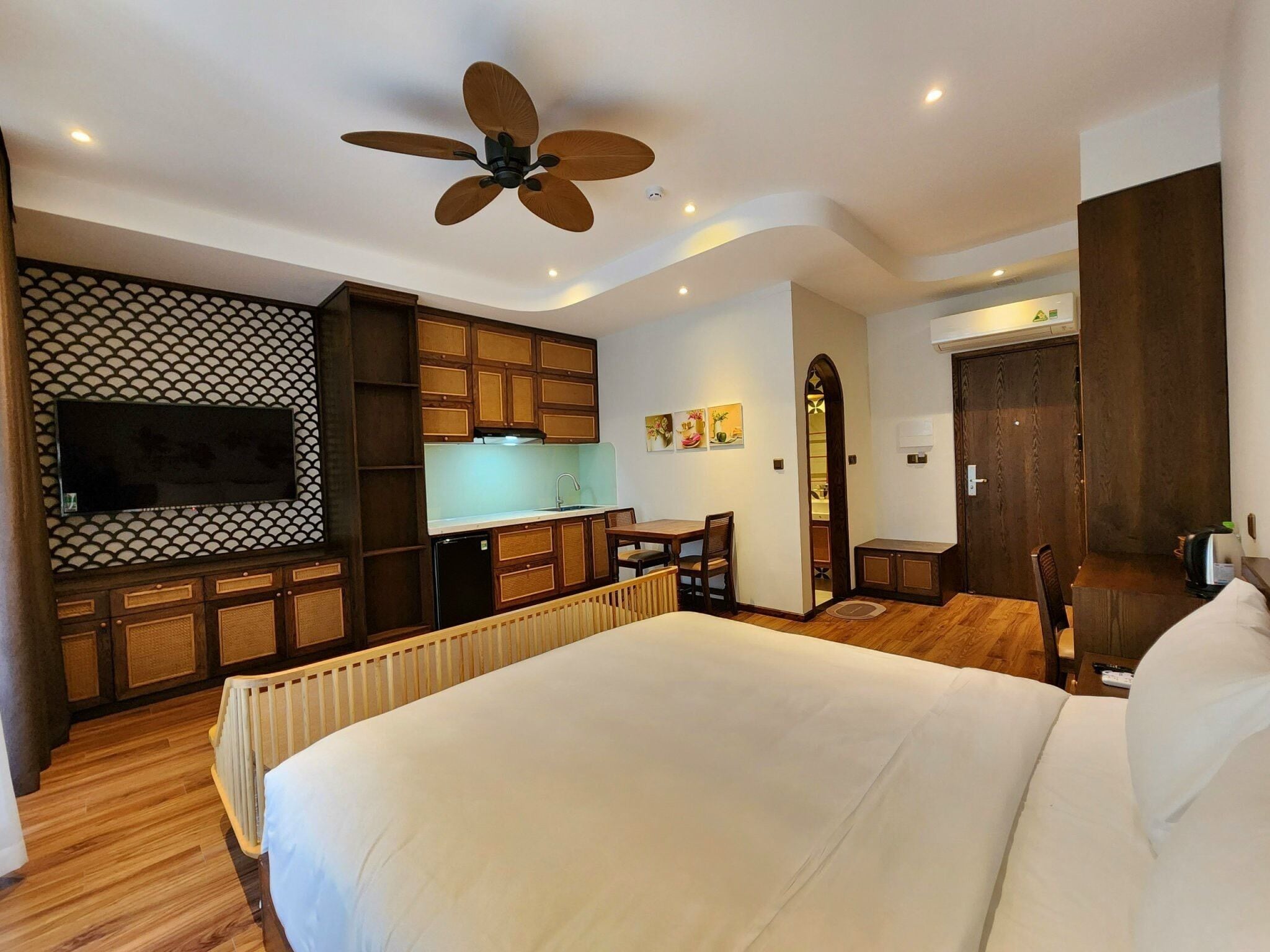 Una habitación de hotel con una cama grande, ventilador de techo, kitchenette y televisión montada en la pared.
