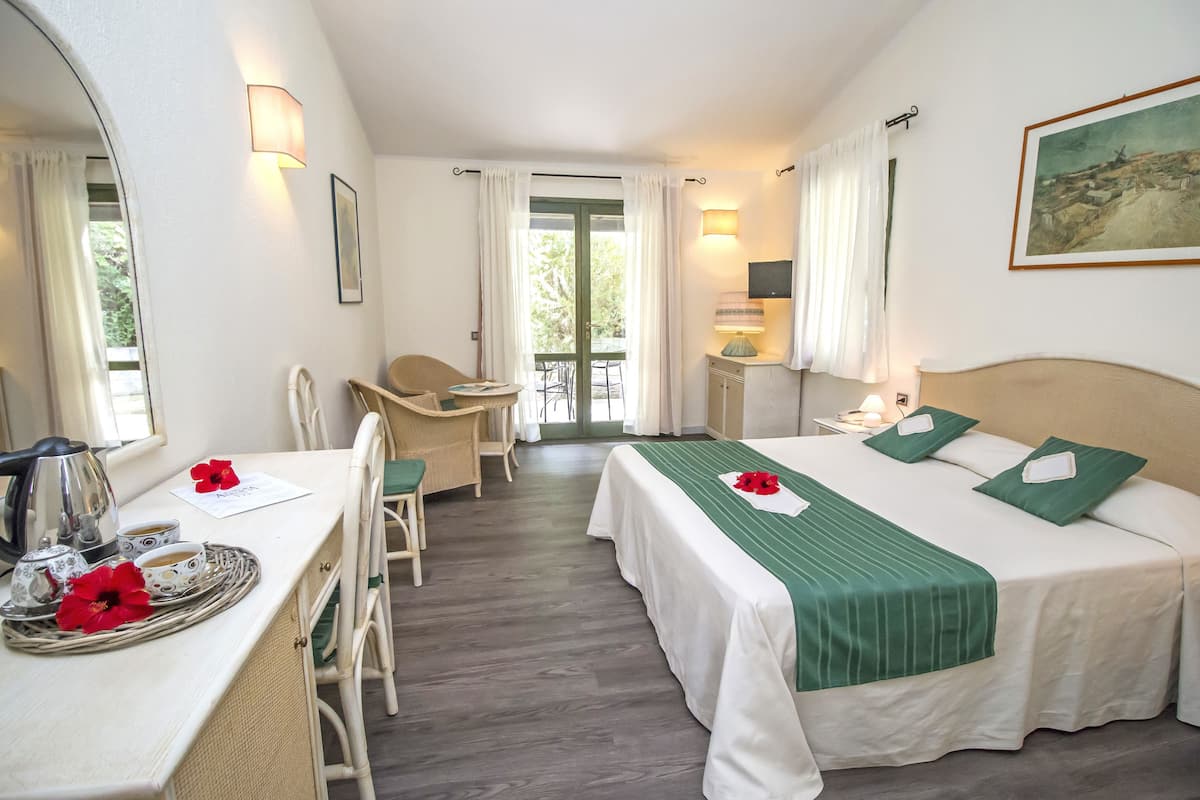 Ett hotellrum med en säng, ett skrivbord med en vattenkokare, koppar och ett bricka med en blomma. Det finns en stol, ett bord och en tavla på väggen.