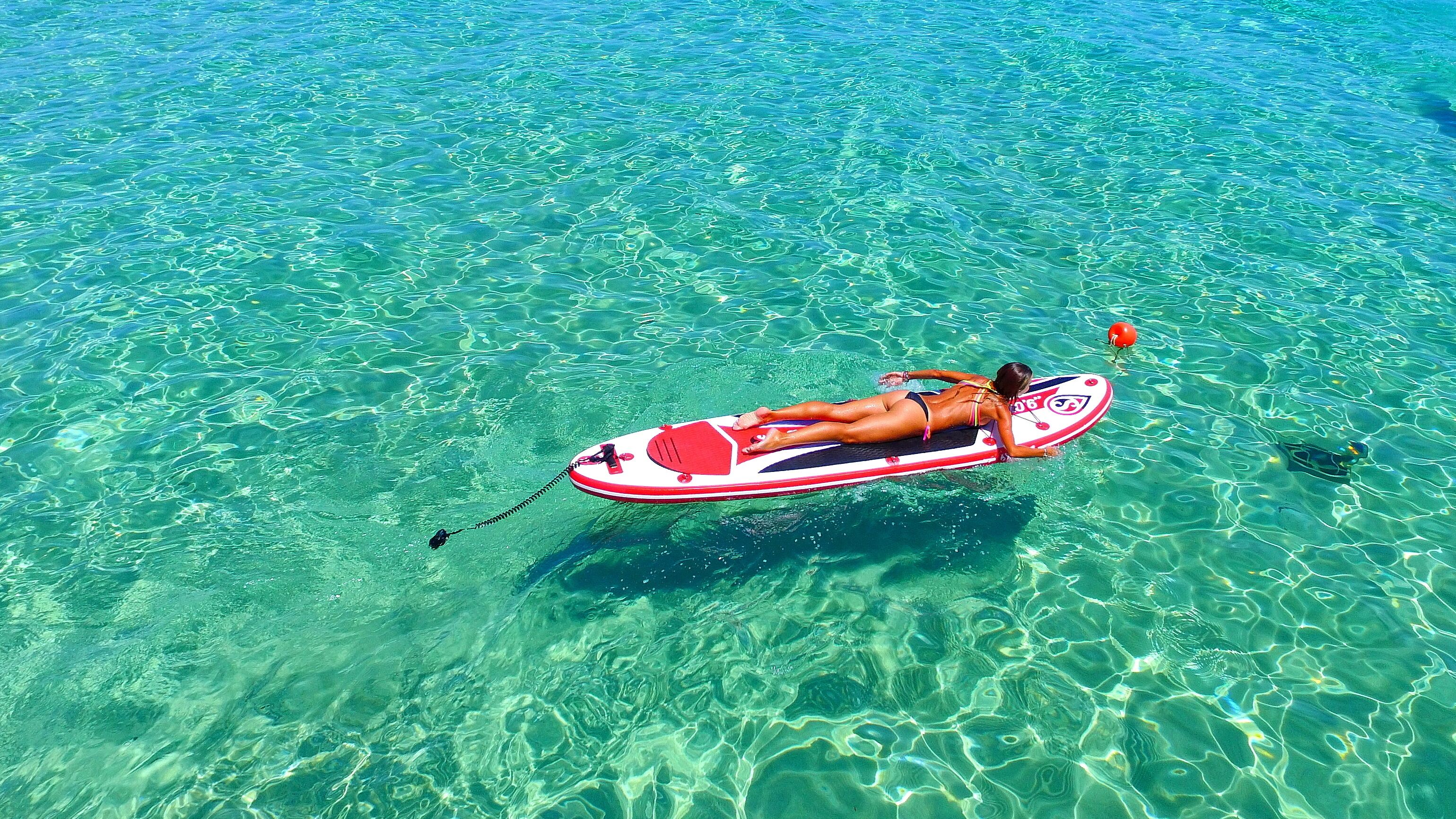 A person lying on a red and white inflatable raft floating on clear turquoise water.