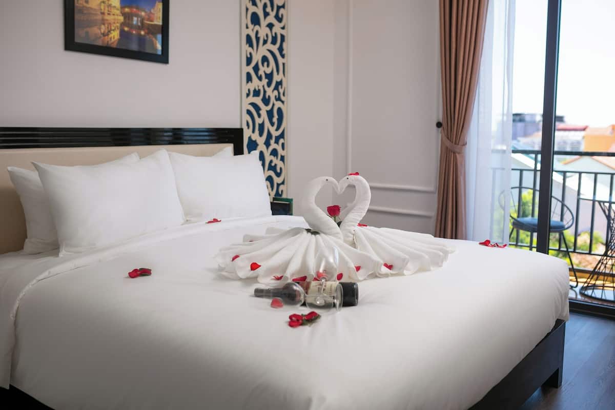 Una cama bien hecha con sábanas blancas, una toalla dispuesta en forma de corazón y pétalos de rosa esparcidos.