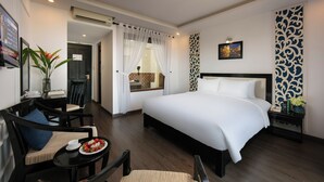 A hotel room with a large bed, a dining area with a table and chairs, a small table with a plate of fruit, and a decorative wall panel.