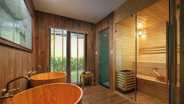 A wooden interior with two wooden bathtubs, a glass door leading to a sauna, and a small table with a bowl.