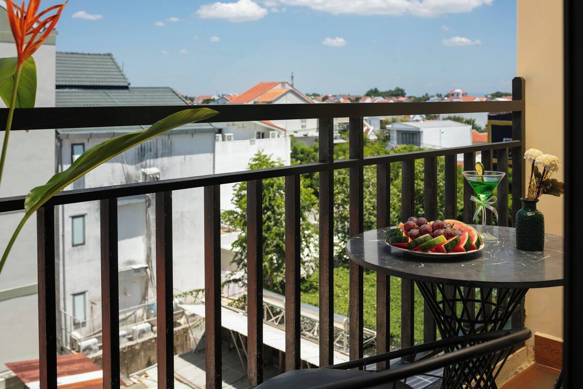 Un balcón con una mesa dispuesta con sandía, uvas y un cóctel verde, con vista a una zona residencial.