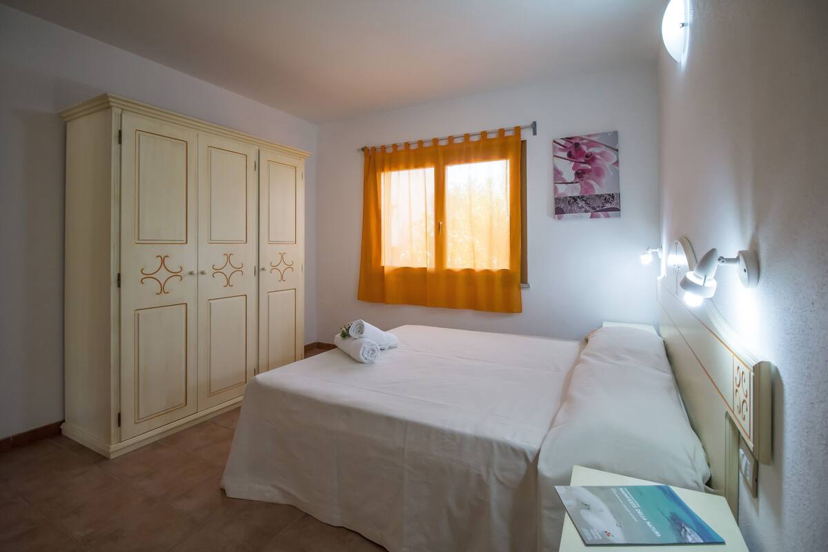 A bedroom with a bed, a wardrobe, a window with curtains, and a wall-mounted light fixture.