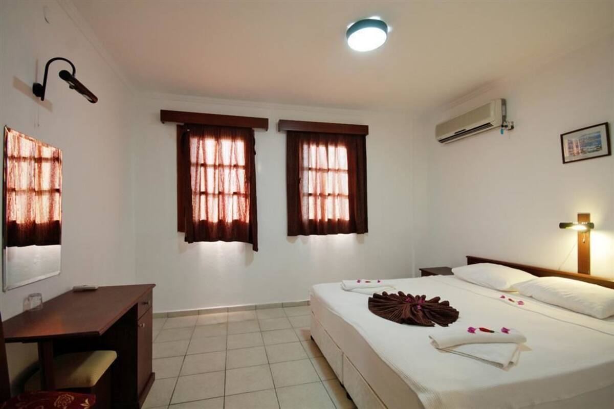 A hotel room with a bed, a desk, two windows with curtains, an air conditioner, and a wall-mounted light fixture.