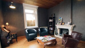 Una sala de estar acogedora con chimenea, dos sillones, una mesa de centro con revistas, una estantería y un ventanal con vistas a árboles.