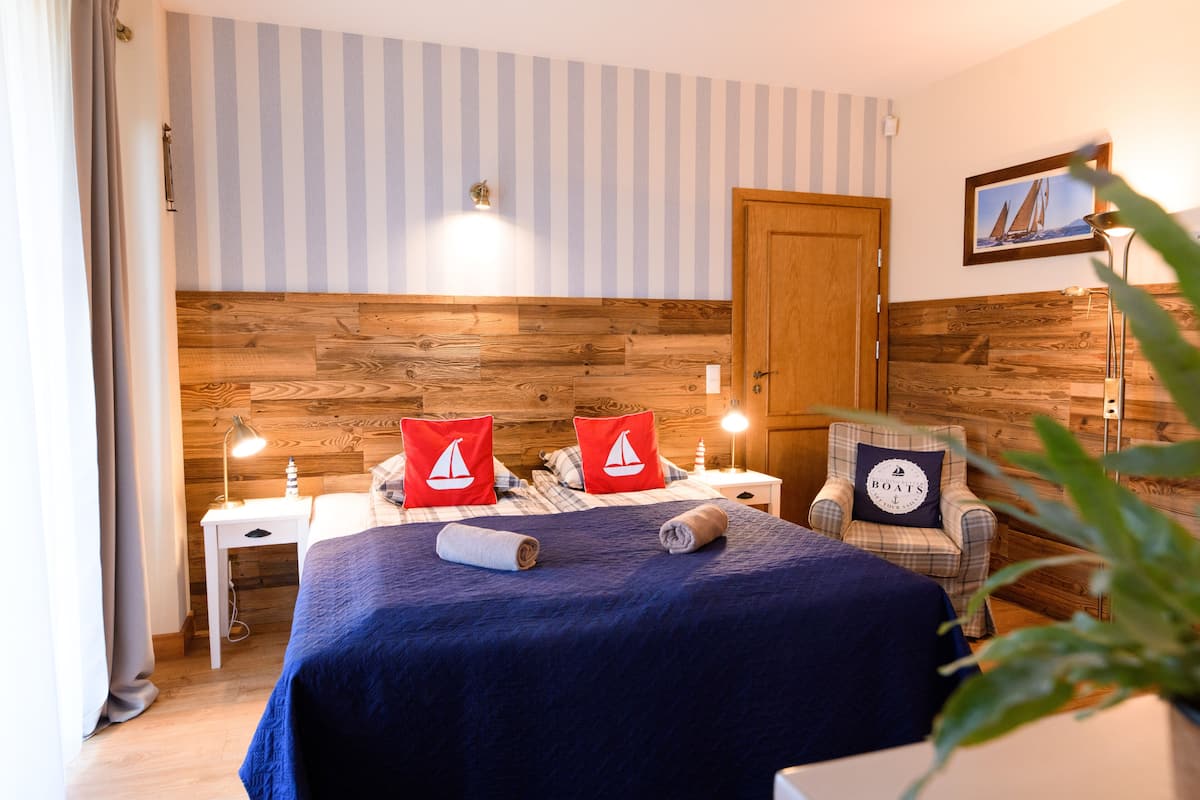 A hotel room with a wooden headboard, striped wallpaper, a bed with blue bedding, two red pillows with sailboat designs, a chair with a pillow labeled "BOATS", and a nightstand with a lamp.