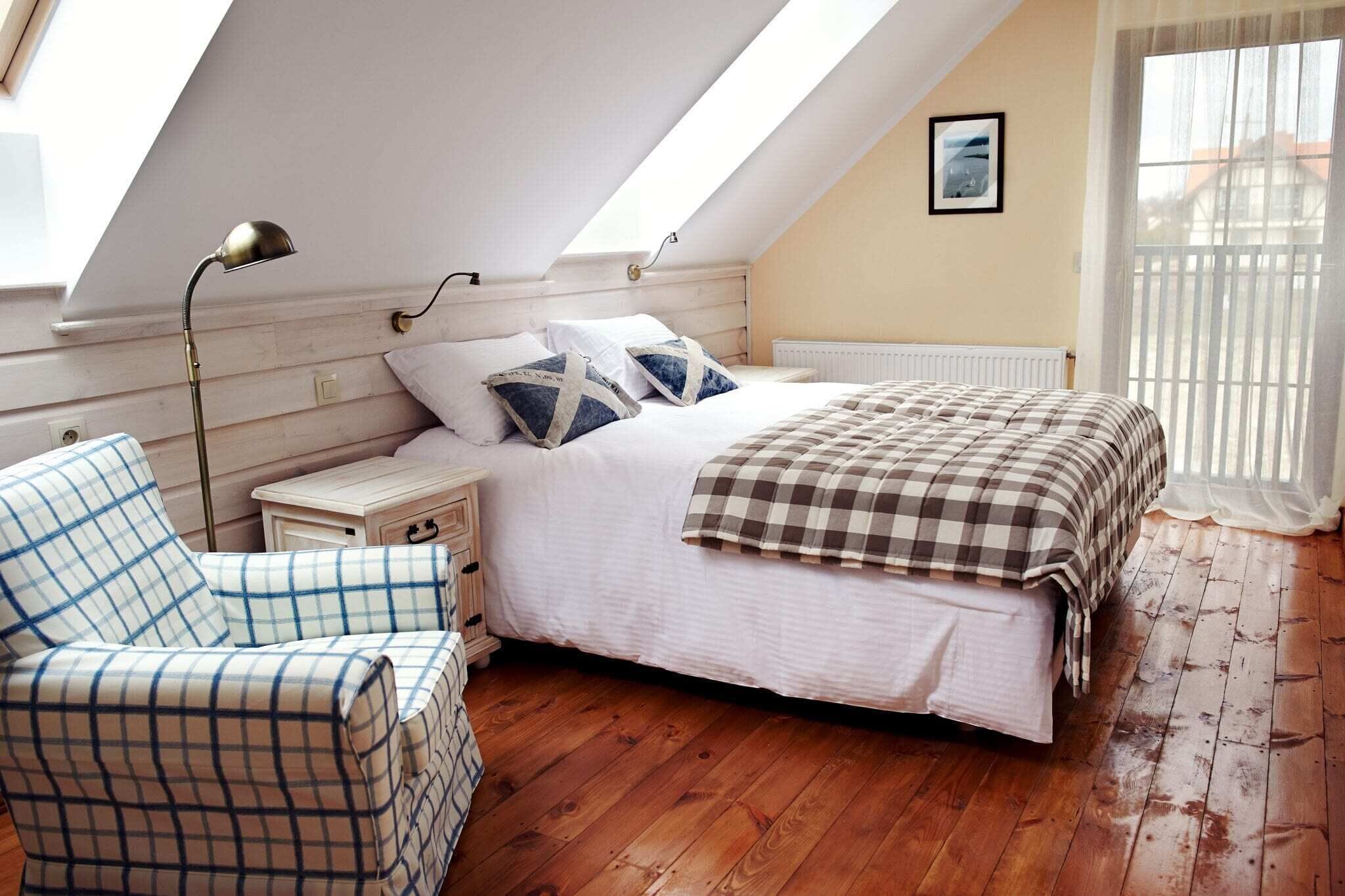 Een slaapkamer met een schuine plafond, een bed met een geruit sprei, een nachtkastje, een stoel en een raam met dunne gordijnen.