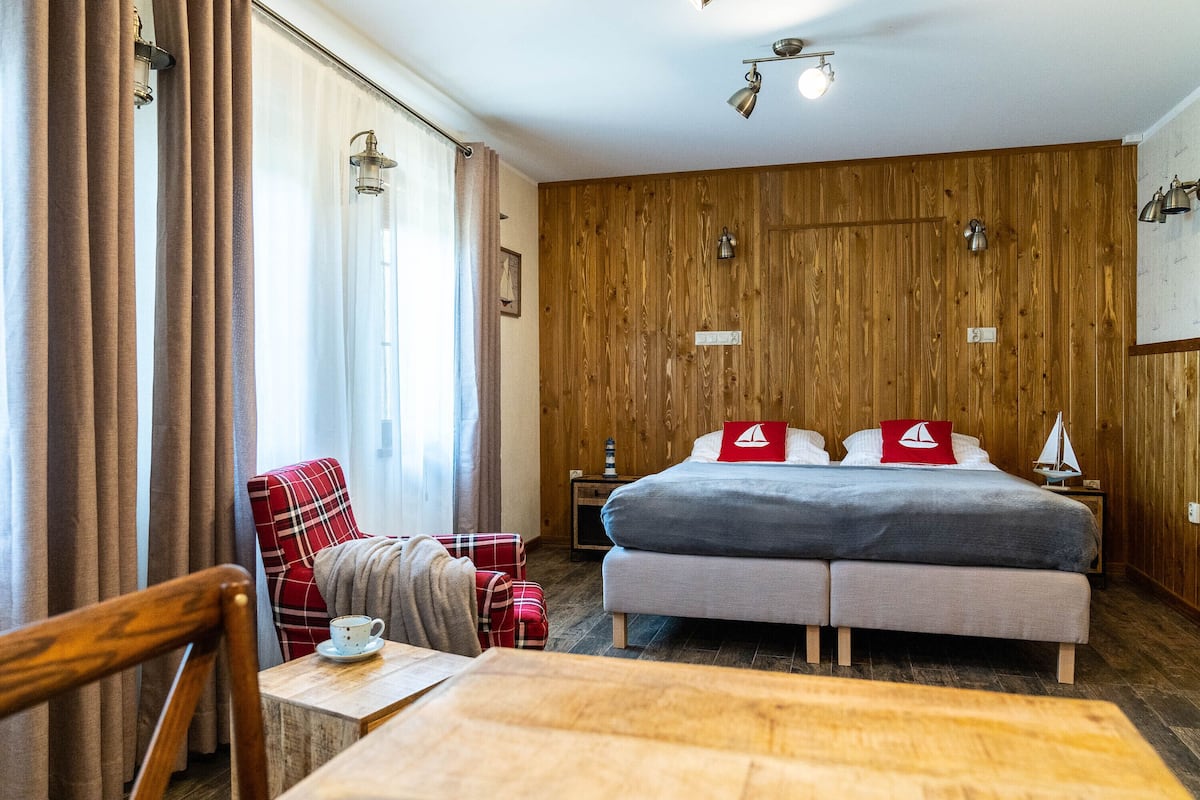A cozy room with a wooden wall, a bed with two pillows, a chair, a small table with a cup, and a nightstand with a lamp.