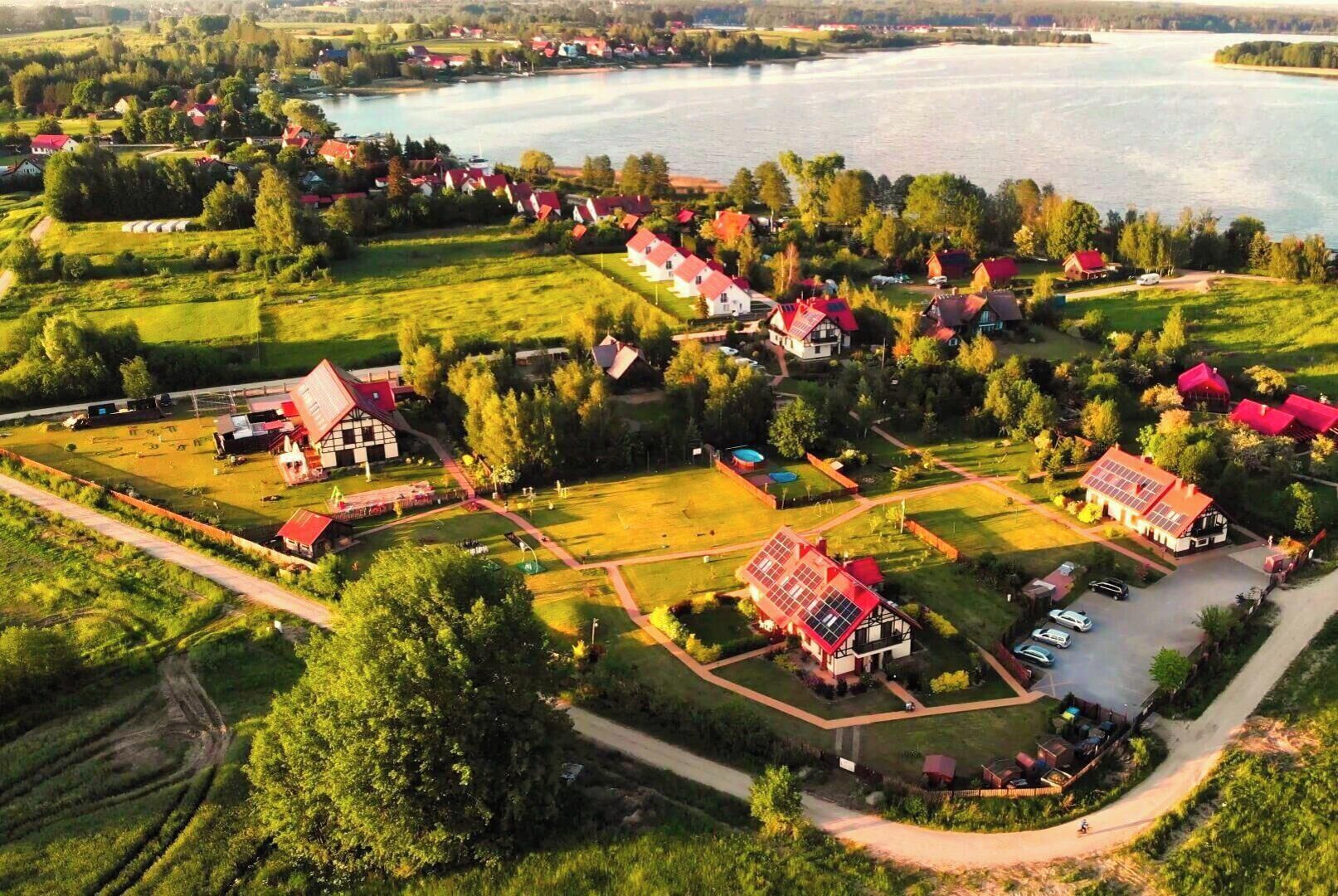 An aerial view of a lakeside village with traditional houses, a swimming pool, and a large barn.