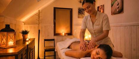 A massage therapy room with a wooden floor, a massage table, a chair, a small table with a candle, and a wall-mounted mirror.