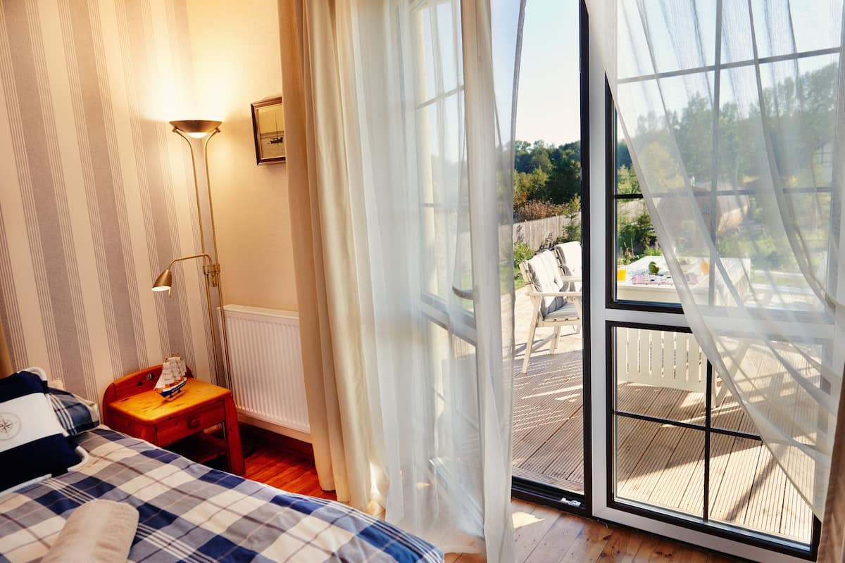 A bedroom with a bed, a nightstand, a lamp, a window with curtains, and a view of an outdoor deck.