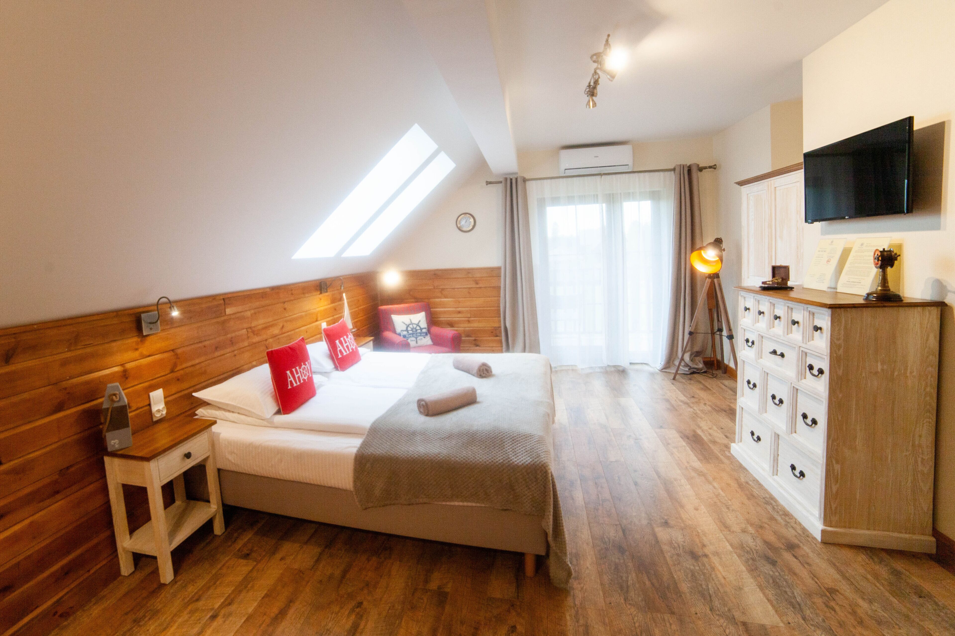 Un dormitorio con cabecera de madera, cama con almohada roja, mesita de noche, cómoda con cajones, televisor de pantalla plana y tragaluz.